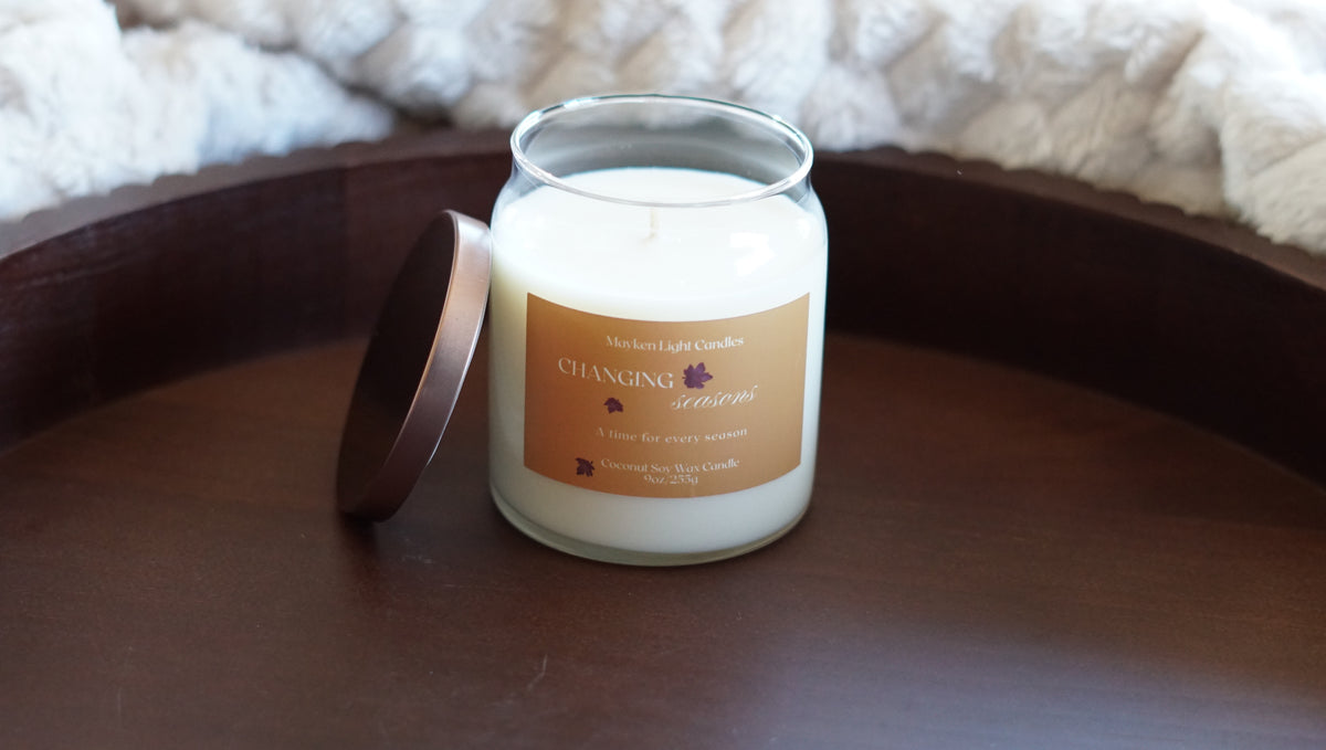 Pictured is a clear candle with a yellow label with the name changing seasons 9 oz candle.