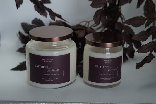 Two white glass jar candles with metallic rose gold lids and dark purple labels, displayed on a light surface with dark leaves in the background.