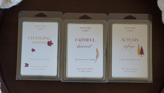 Three Mayken Light Candles soy wax melts in clear plastic clamshell packaging, displayed side-by-side on a dark brown wooden surface. The packages feature white labels with elegant typography and autumn-themed illustrations: 'Changing Seasons' with red leaves, 'Faithful Harvest' with a wheat stalk, and 'Autumn Refuge' with pine trees.