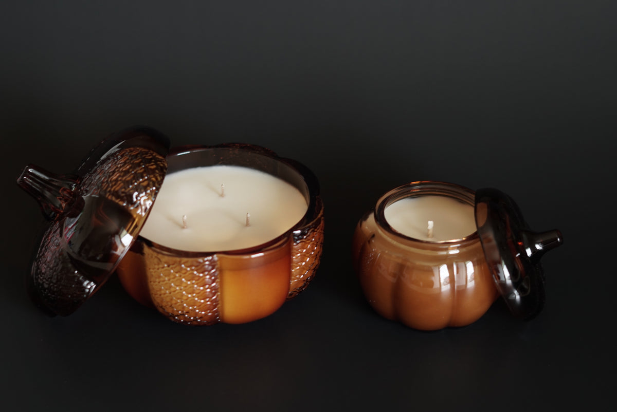 2 Amber Glass Pumpkin Candles. Lids leaned on the side of each jar with a black background 