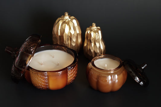 One large amber glass pumpkin with 3 weeks and 1 mini amber glass pumpkin with 1 wick. 2 gold pumpkins in background. 