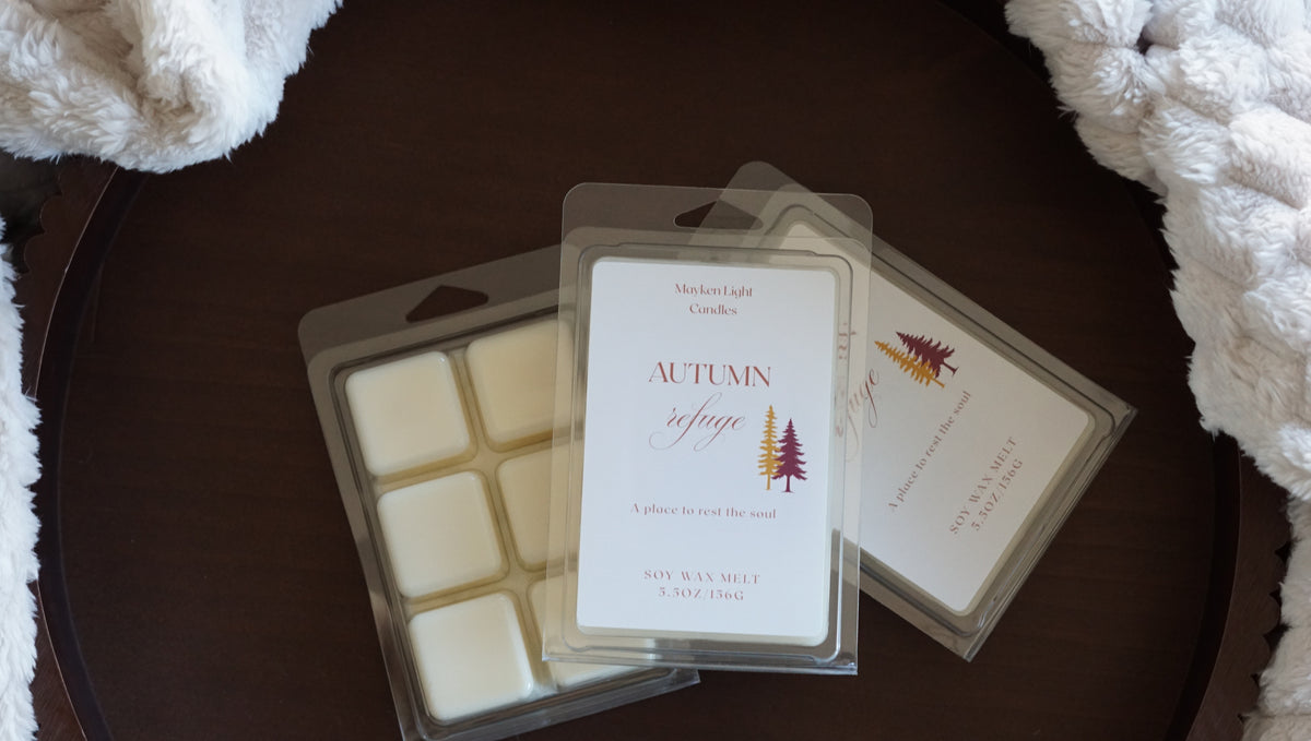 Pictured is a clear container on a brown tray and a plush blanket with the name autumn refuge wax melt 5.5 oz. 
