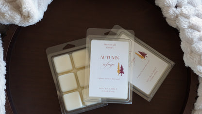 Pictured is a clear container on a brown tray and a plush blanket with the name autumn refuge wax melt 5.5 oz. 
