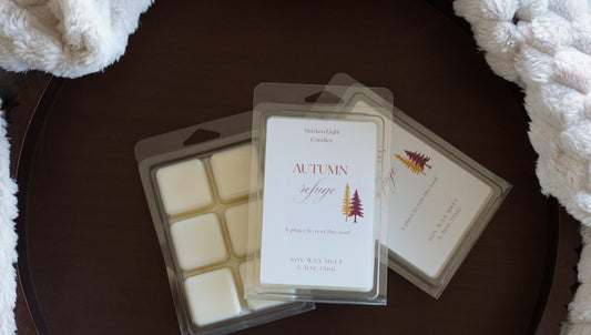 Pictured is a clear container on a brown tray and a plush blanket with the name autumn refuge wax melt 5.5 oz. 