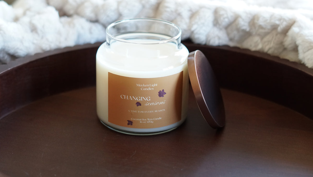 Pictured is a candle with a yellow label that says changing seasons, 16 oz candle on a wooden tray and a plush quilt in the background 