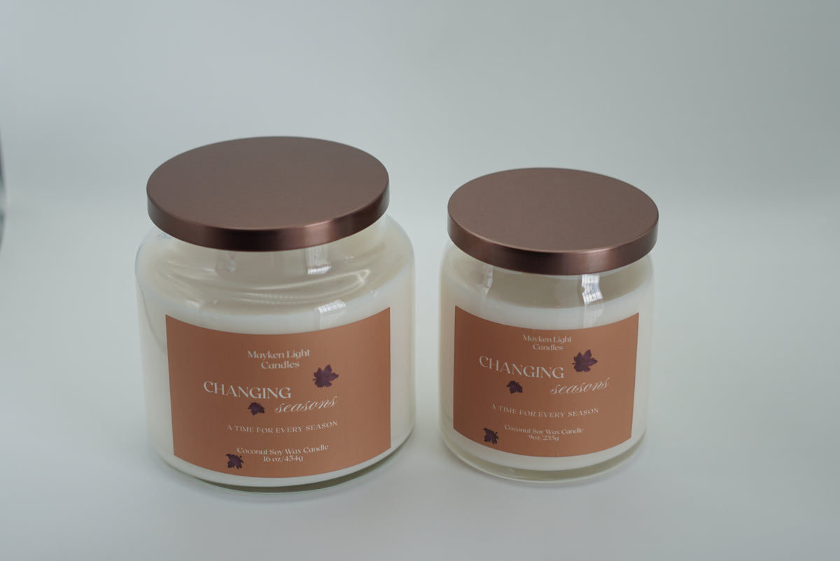 Two large glass jar candles with cream-colored wax and Yellow labels, one open with its lid leaning beside it and one closed with a metallic rose-gold lid, set against a dark background.