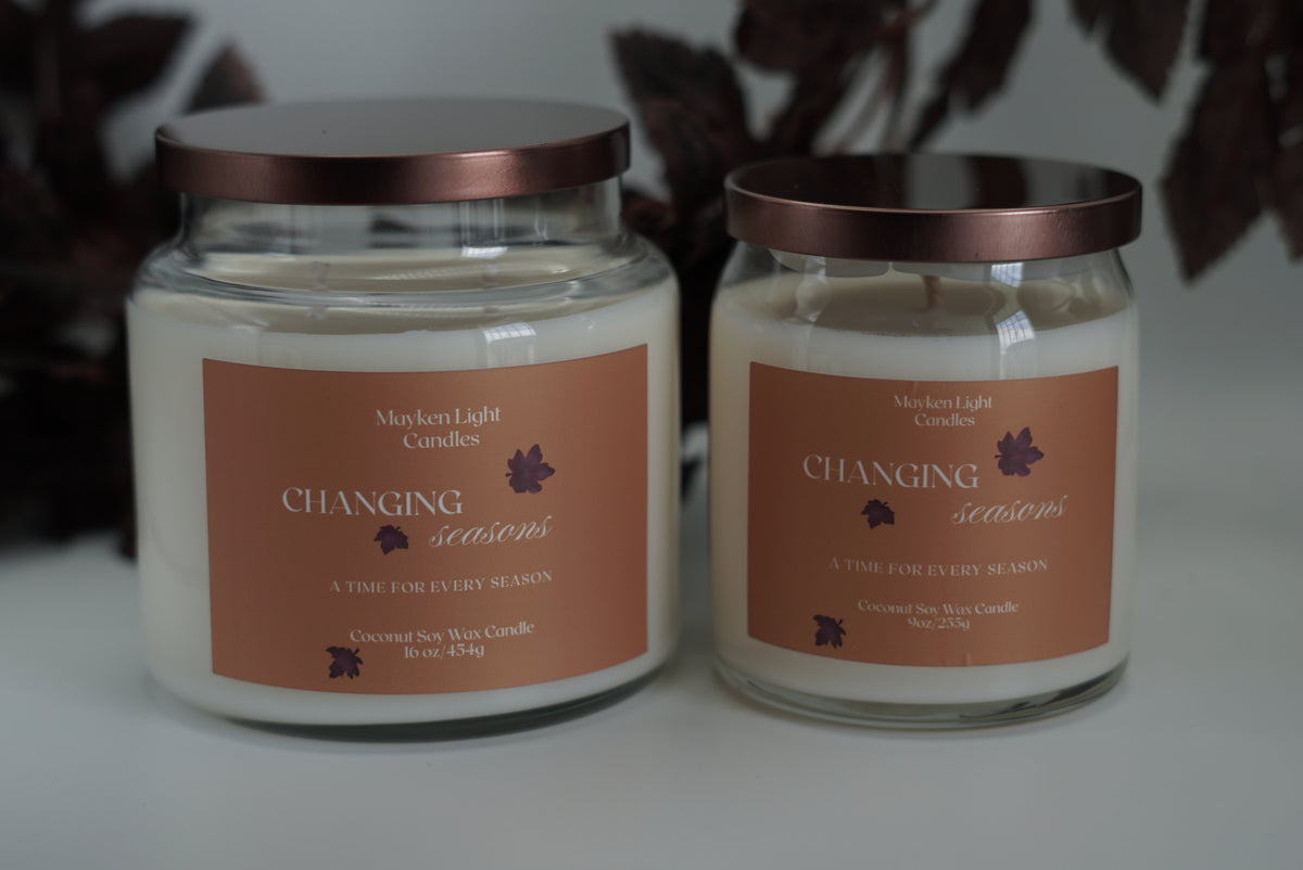 Two large glass jar candles with cream-colored wax and Yellow labels, one open with its lid leaning beside it and one closed with a metallic rose-gold lid, set against a dark background.