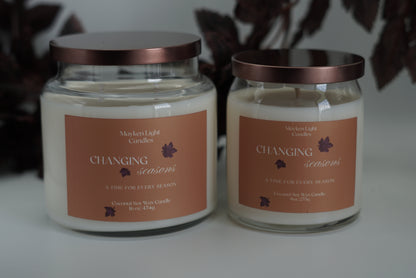 Two large glass jar candles with cream-colored wax and Yellow labels, one open with its lid leaning beside it and one closed with a metallic rose-gold lid, set against a dark background.