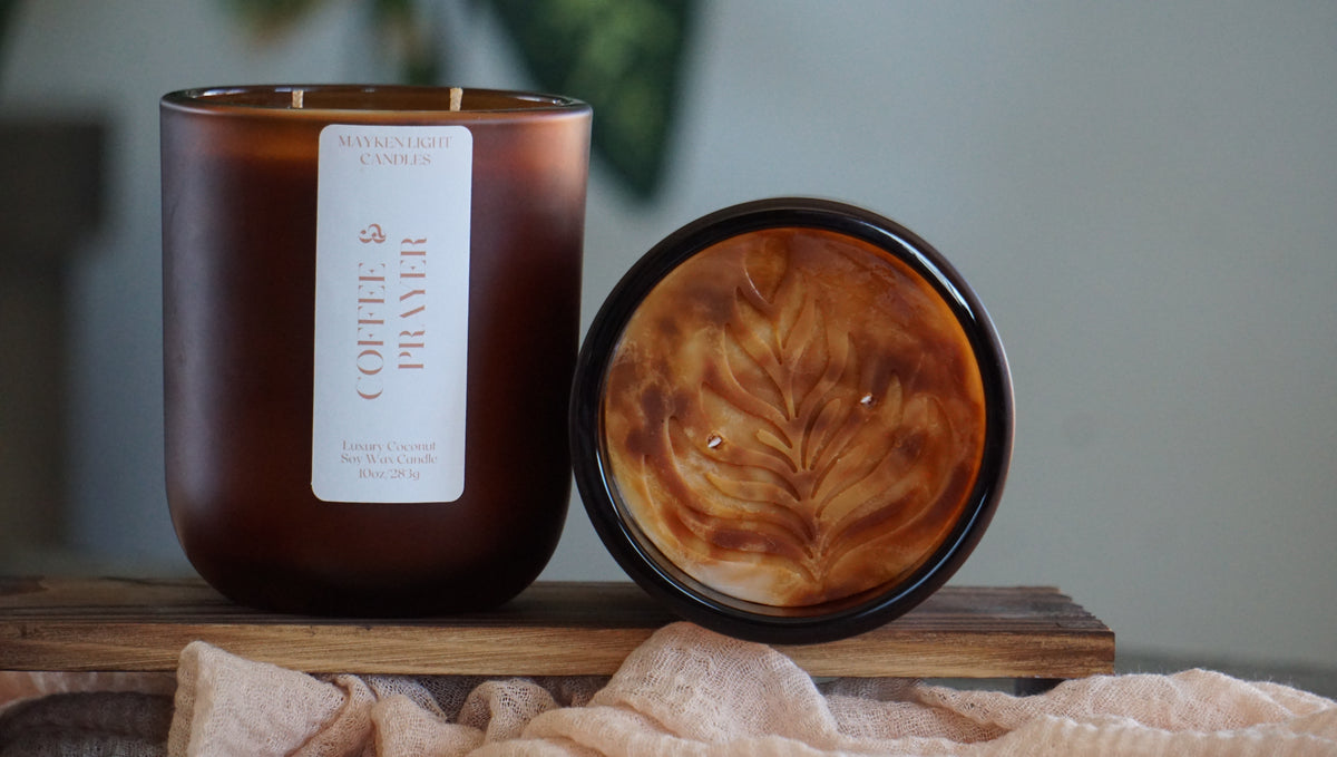 Two brown candle jars. One showing white label reading Coffee & Prayer. The other showing a Rosetta Latte art. Both on a brown shelf with light tan material in front and a green plant in back. 