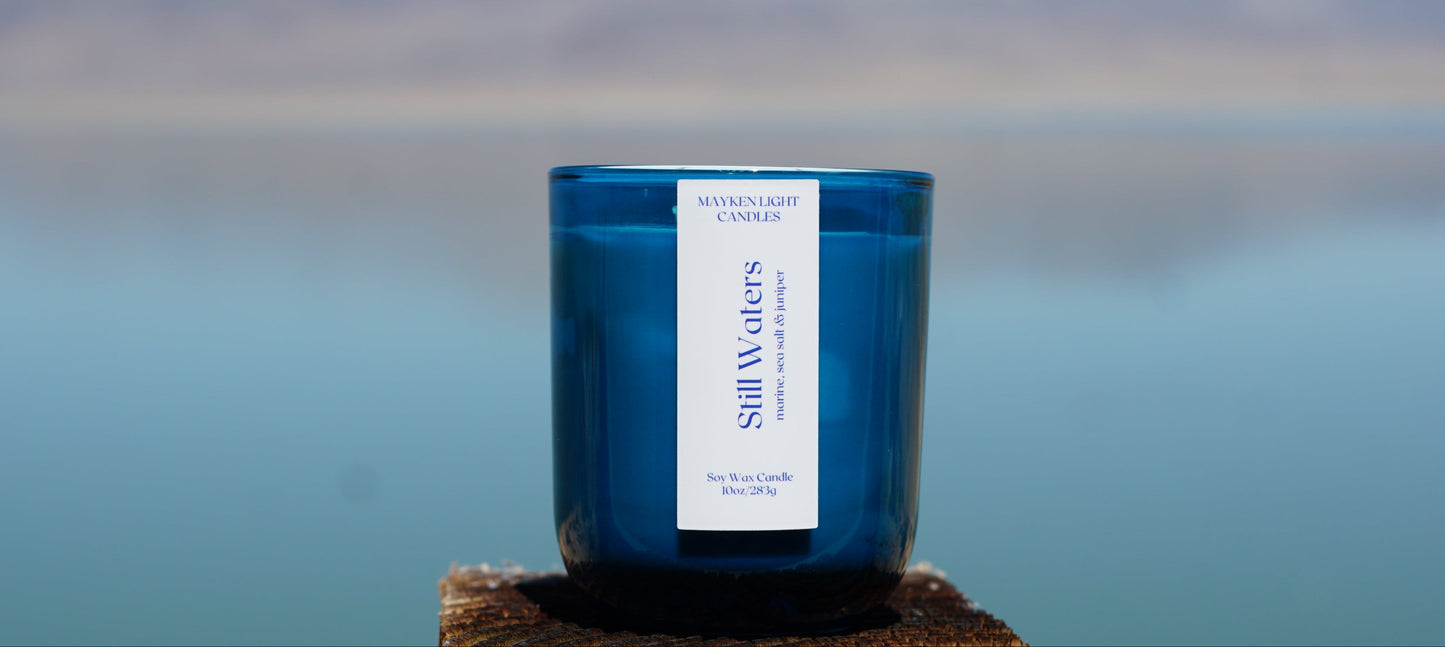A blue glass jar candle labeled 'Still Waters' by Moonlight Candles, placed on a wooden surface with a blurred ocean and sky background.