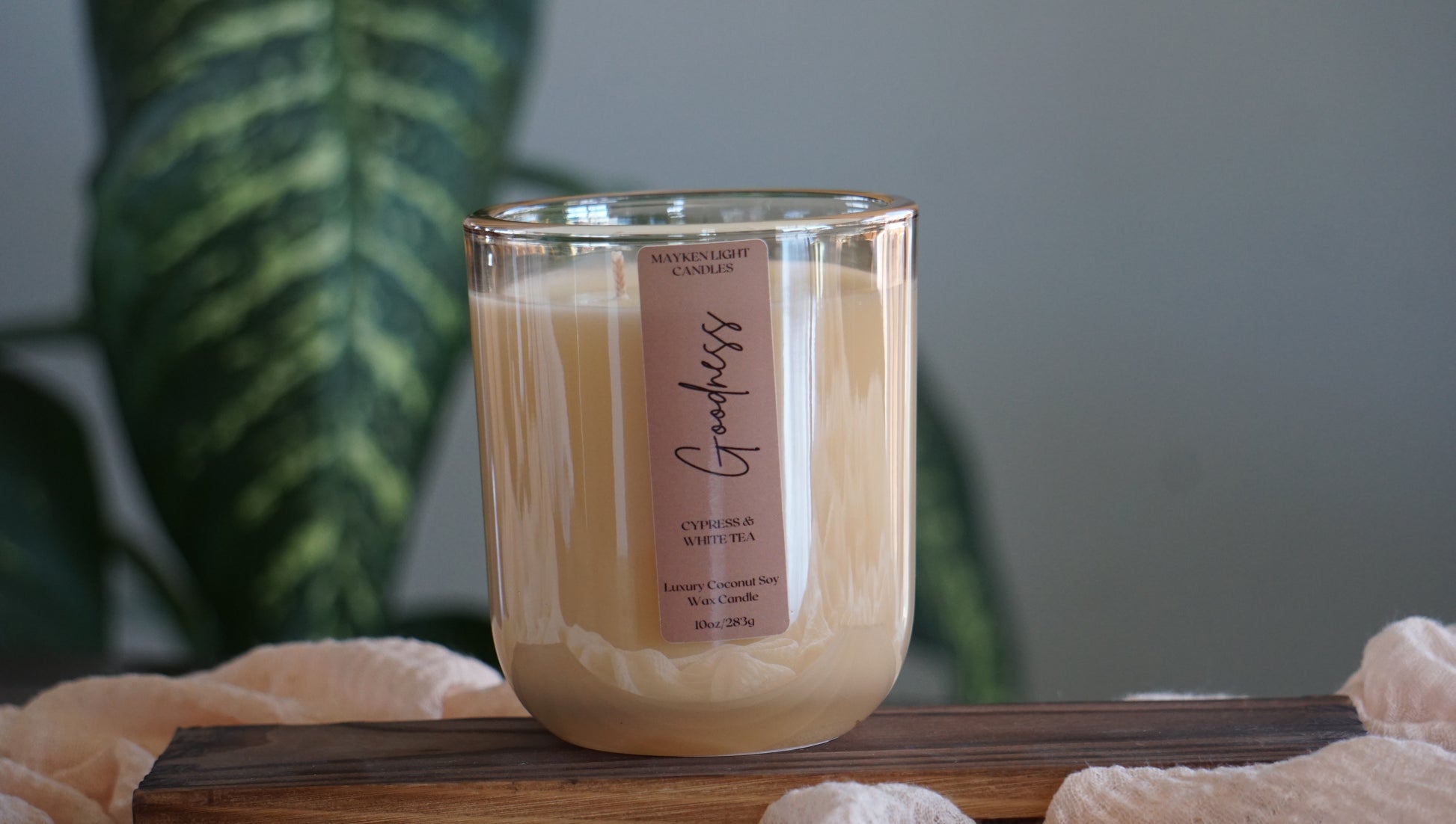 Candle with a label on a wooden surface, blurred green plant in the background