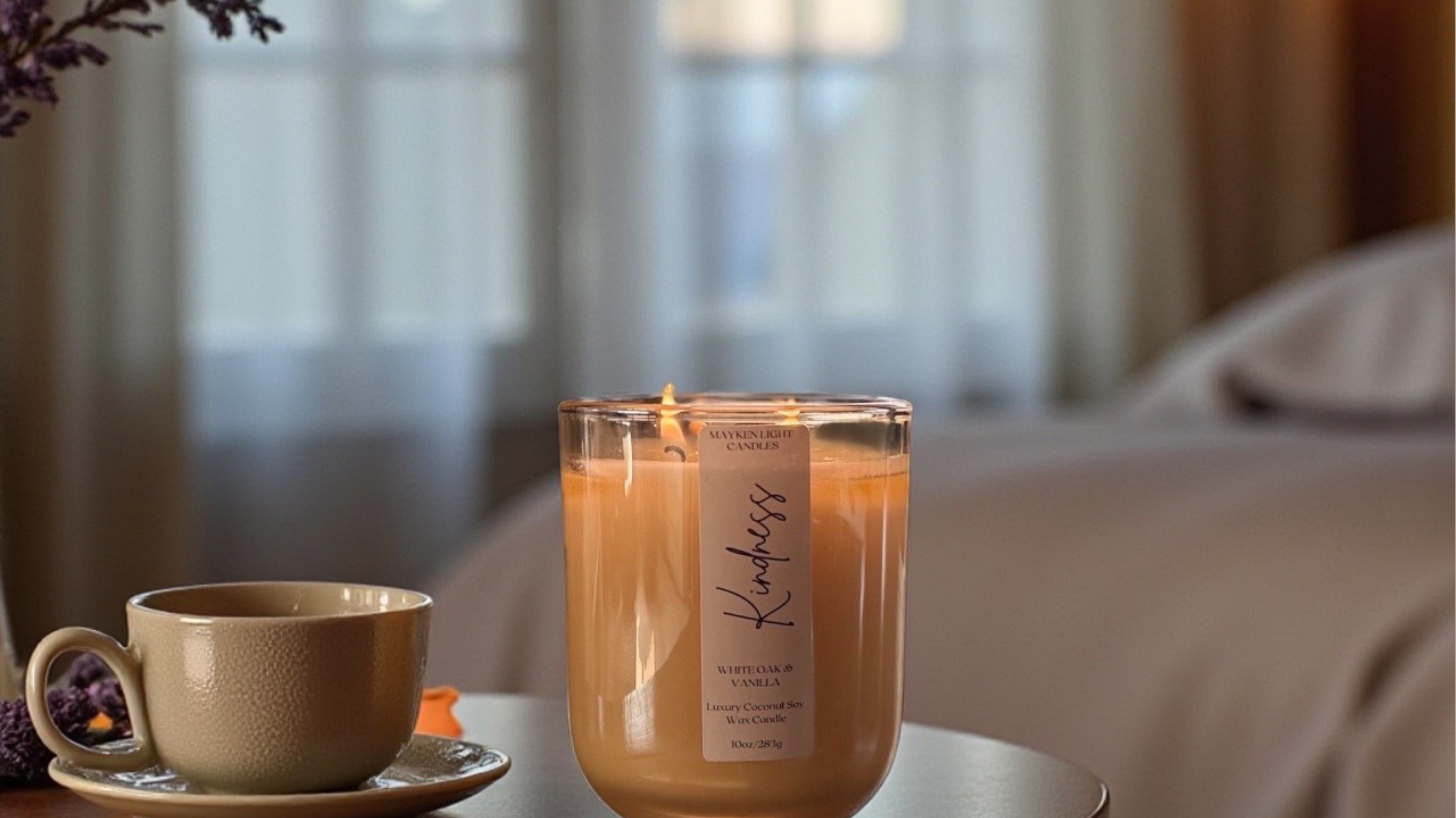 Candle labeled 'Kindness' with a cup on a table in a softly blurred room setting
