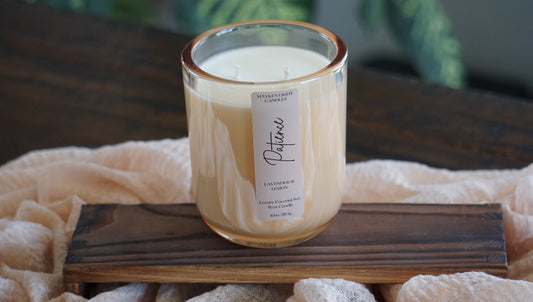 Candle with a label on a wooden surface with soft material around it and a green plant in ackground