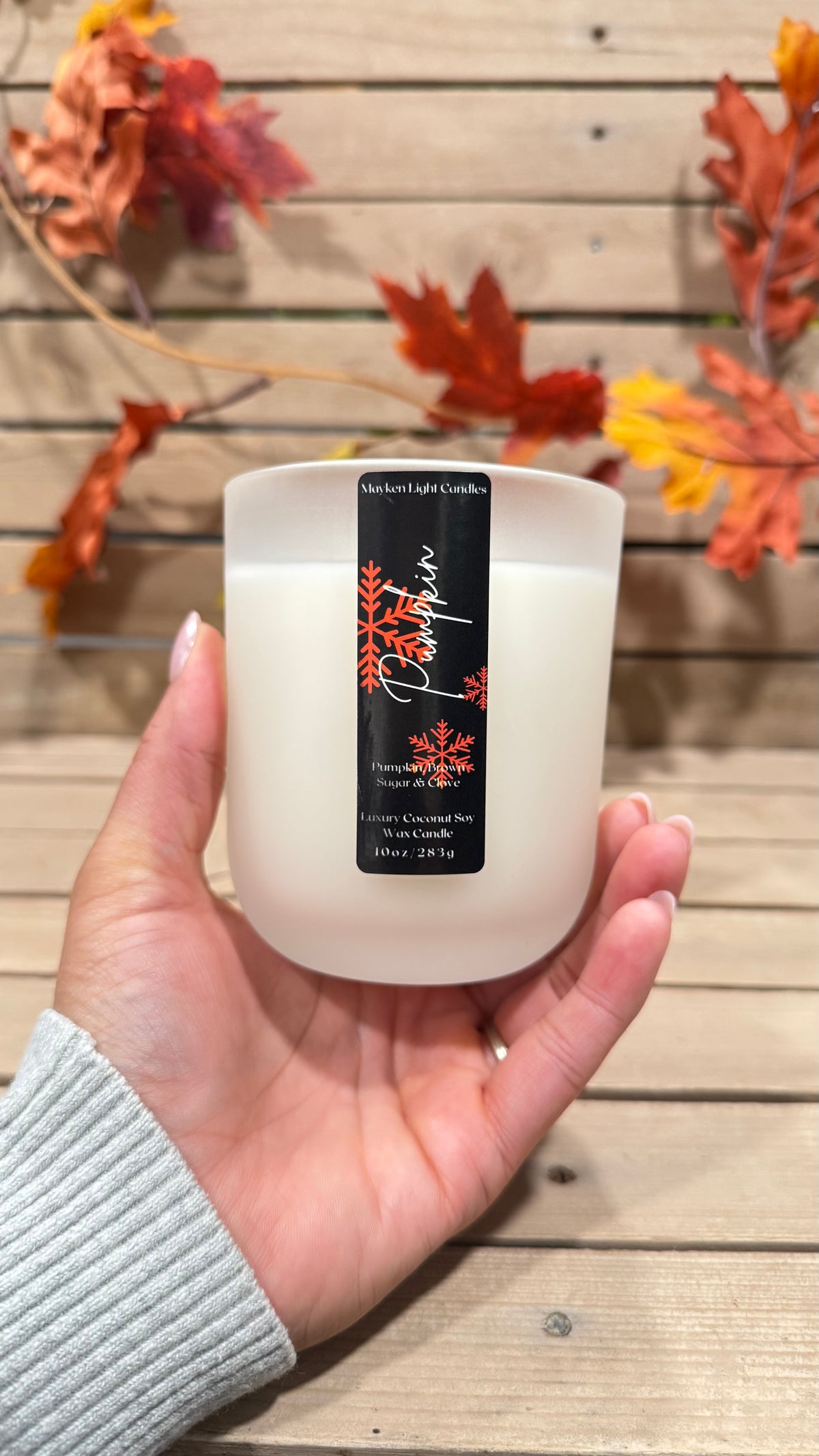 White Candle with a black label that reads Pumpkin in and has orange snowflakes held up with a hand in front of a wooden fence and fall leaves. 