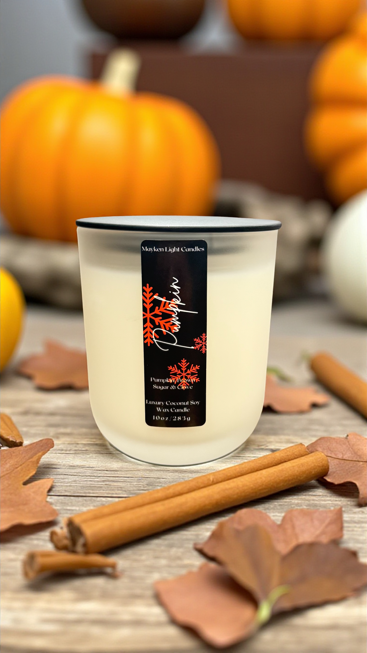 White Candle with a black label that reads Pumpkin and has orange snowflakes set in front of decorative pumpkins, brown leaves and cinnamon sticks on a wooden surface 